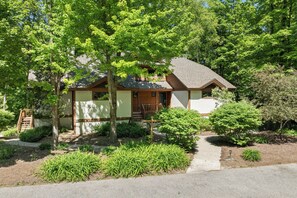 A single-story house with a front porch, surrounded by mature trees and a well-maintained garden. - Disciples Village #377 Condo | Walking Distance to the Slopes! (Boyne Falls)