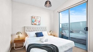 A modern hotel room with a large bed, a glass door leading to a balcony, and a colorful abstract painting on the wall.