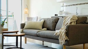 A modern living room with a grey sofa, a round wooden coffee table, and a small side table. A kitchen with white cabinets and a tiled backsplash is visible in the background.