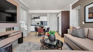 A modern living room with a sofa, a coffee table, and a TV. There is a dining area with chairs and a table. The kitchen is visible in the background. - Modern 2-bedroom Executive apartment with WiFi, in Masingita Towers Sandton (Sandton)