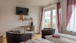 Ein Hotelzimmer mit Bett, zwei Sesseln, einem Schreibtisch mit Stuhl, einem Flachbildfernseher und einem Fenster mit Vorhängen.