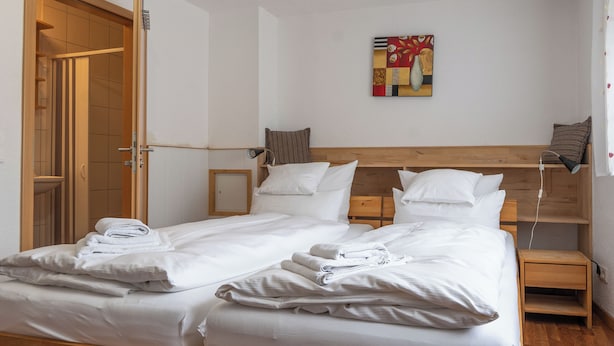 A double bed with white linens and towels, a wooden headboard, a bedside table with a lamp, and a small shelf. - Gästehaus Neckarsulm (Neckarsulm)