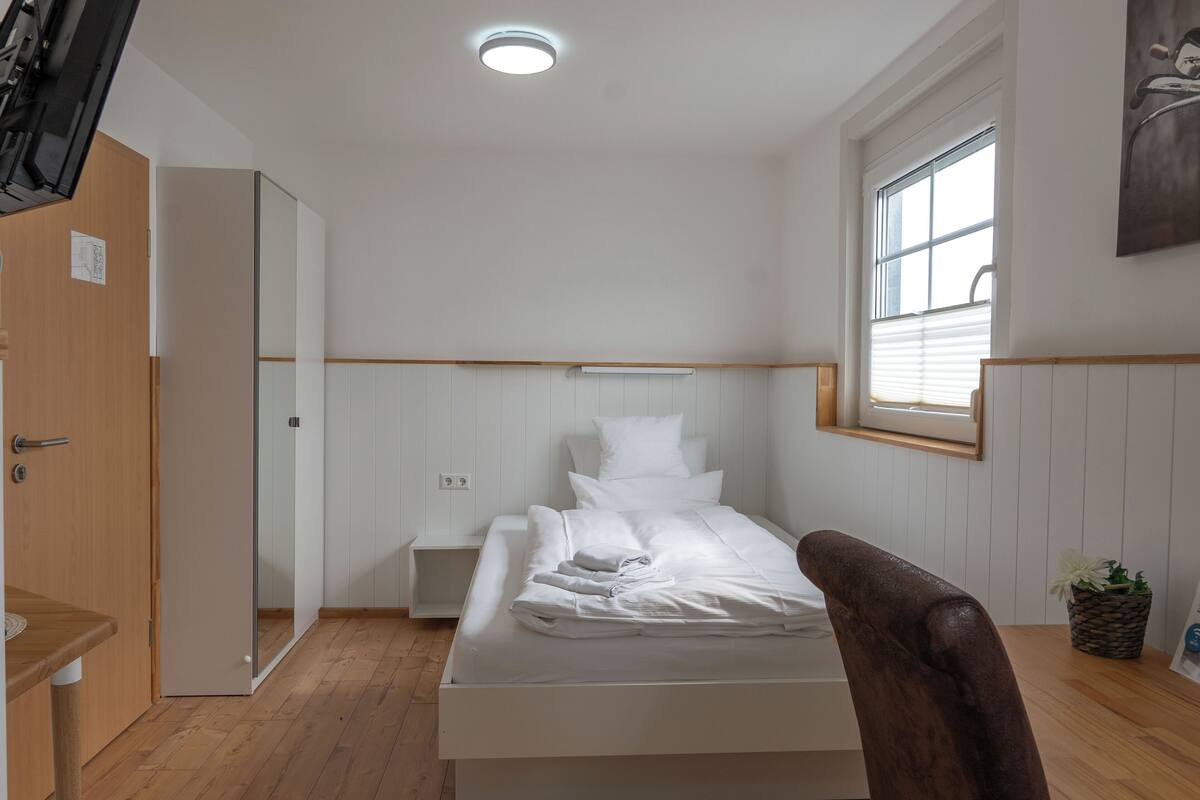 A small, clean room with a bed, a wooden chair, a small table, and a window with blinds.
