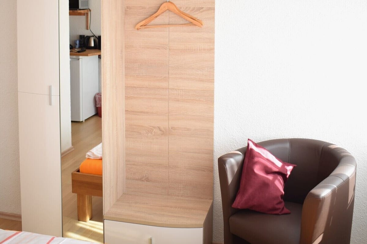 A bedroom with a wardrobe, a bed, a chair, and a hanging coat rack.