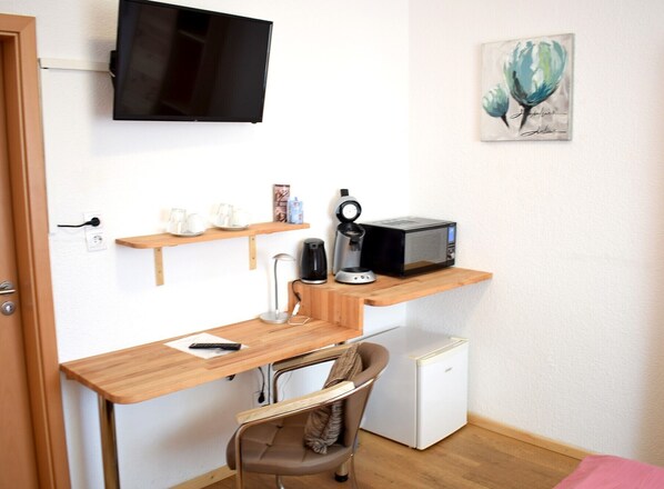 A compact room with a wooden desk, a small refrigerator, a microwave, a coffee maker, and a TV mounted on the wall. - Gästehaus Neckarsulm (Neckarsulm)