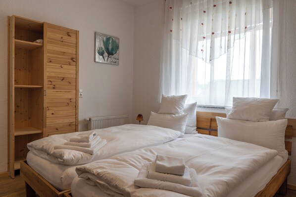 Un lit double avec du linge de lit blanc, une armoire en bois et une fenêtre ornée de rideaux légers.