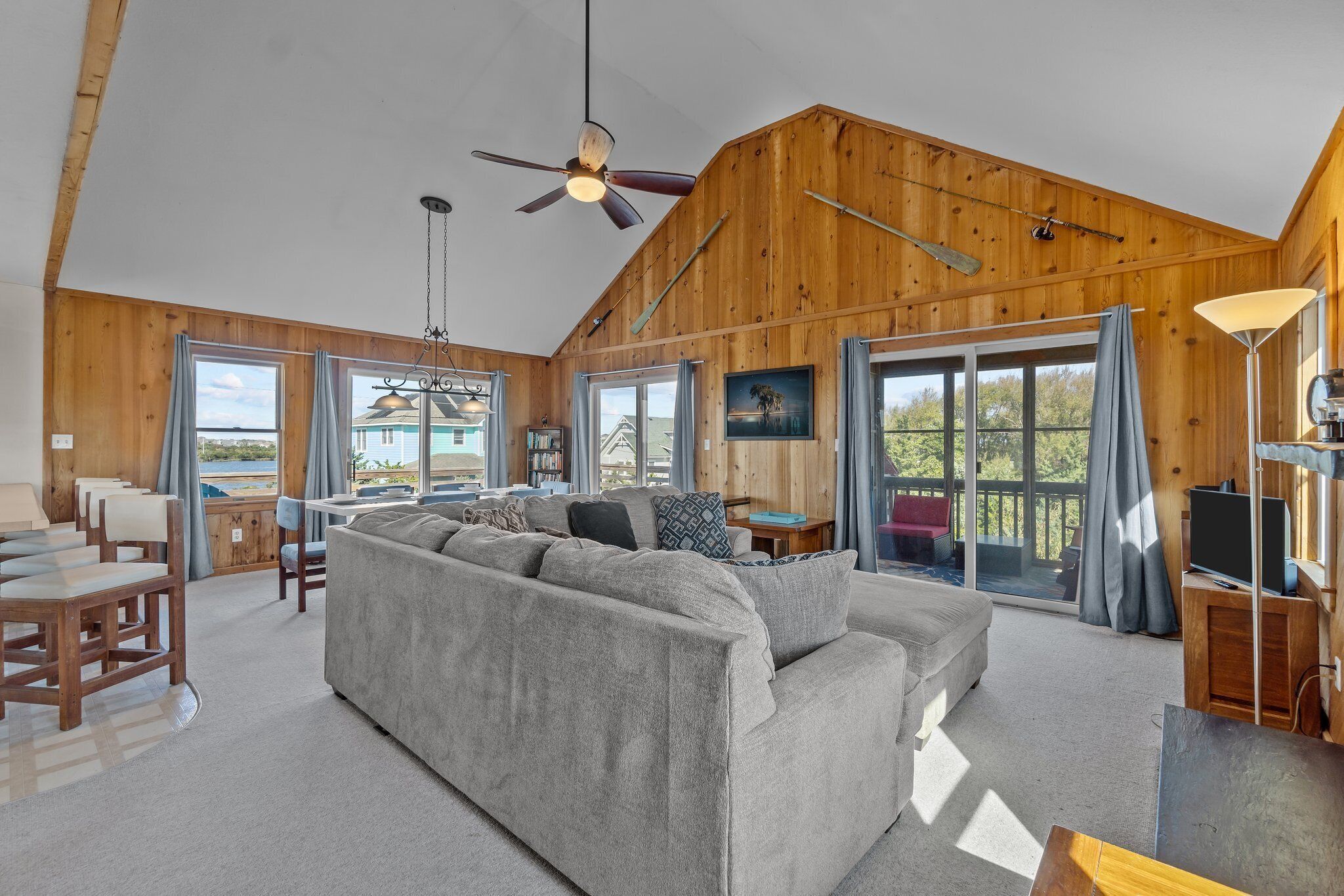 A spacious living room with a large gray sofa, wooden walls, and large windows offering a view of the outdoors.