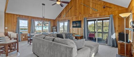 A spacious living room with a large gray sofa, wooden walls, and large windows offering a view of the outdoors.