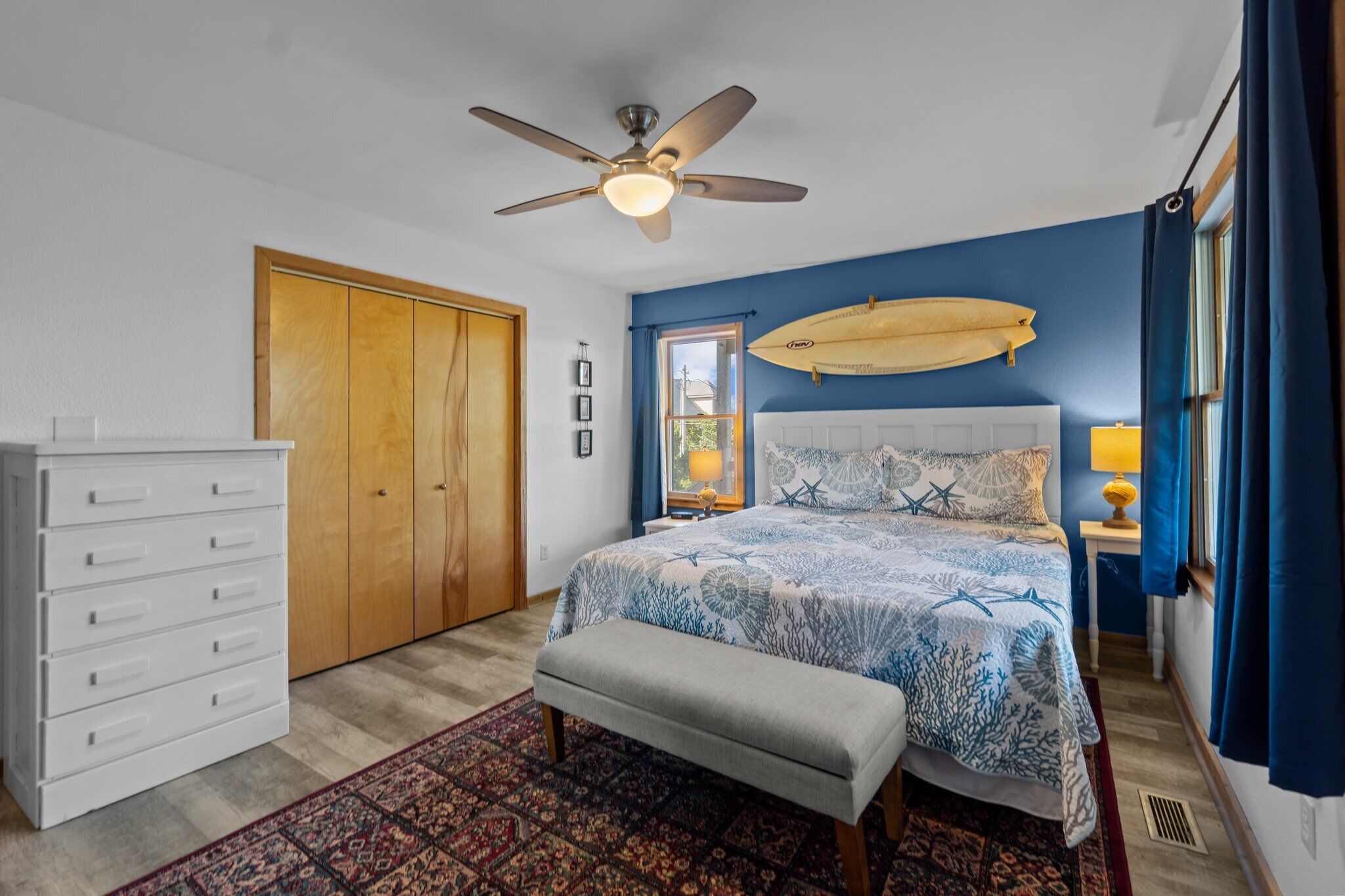 Un dormitorio con cama, ventilador de techo, armario de madera, banco y ventana con cortinas.