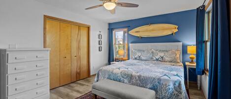 Un dormitorio con cama, ventilador de techo, armario de madera, banco y ventana con cortinas.