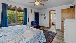 A bedroom with a large bed, a ceiling fan, a window with curtains, a chair, and a view of the outside. - Prime Location, Short Walk To The Beach, Water Views, Sun Deck (Rodanthe)