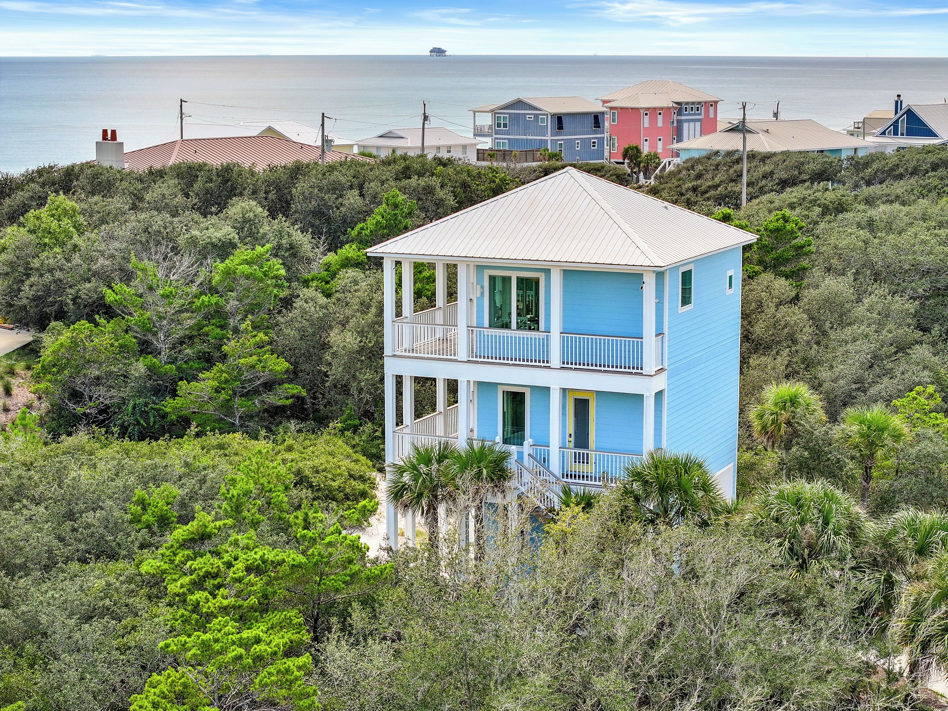 Una casa costera de dos pisos con balcón, rodeada de abundante vegetación y vista al mar.