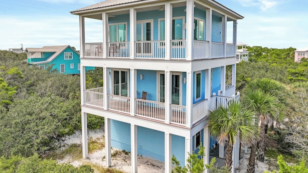 Casa de playa de dos pisos con varios balcones, rodeada de vegetación exuberante y otros edificios.
