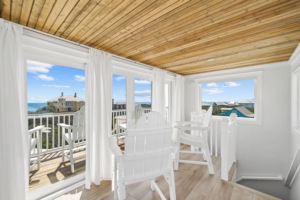Un balcon abrité, doté de chaises blanches et d’un plafond en bois, qui offre une vue sur un quartier résidentiel avec ses maisons et un ciel dégagé.