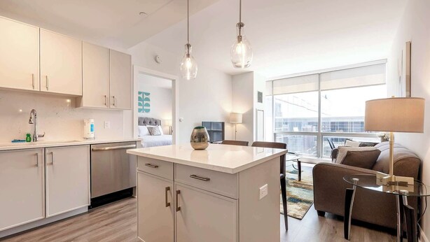 A modern kitchen with white cabinets, a central island, and pendant lighting. There is a dining area with a sofa and a lamp. - Luxury Apt Near Downtown (Stamford)