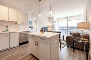 A modern kitchen with white cabinets, a central island, and pendant lighting. There is a dining area with a sofa and a lamp. - Luxury Apt Near Downtown (Stamford)