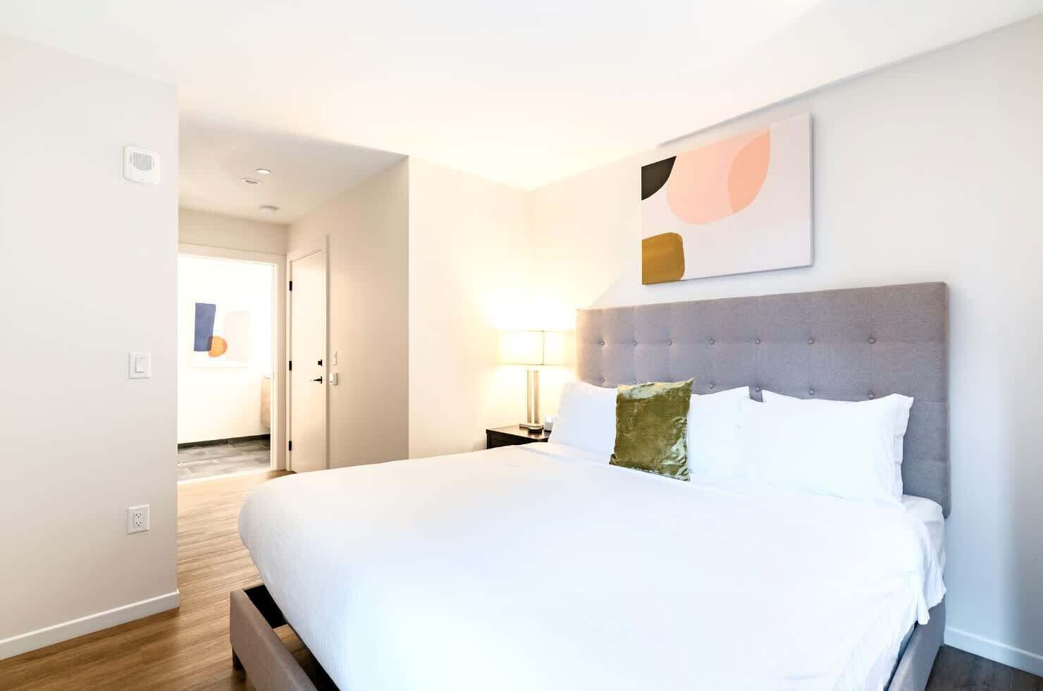 A modern hotel room with a large bed, a grey upholstered headboard, and a wall-mounted abstract artwork.