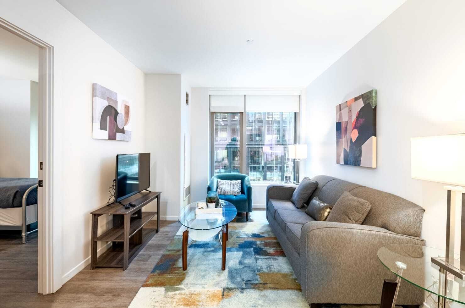 Un salon moderne avec un canapé, une table basse en verre, un téléviseur et une vue sur la ville grâce à de grandes fenêtres.
