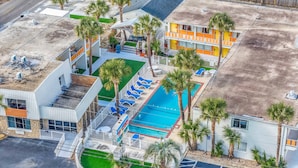 An aerial view of a hotel with a swimming pool, palm trees, and a parking lot.
