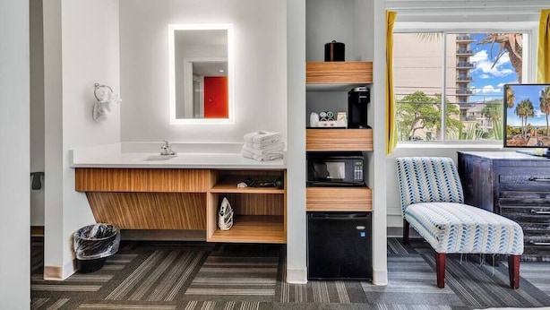 A modern bathroom with a white vanity, a mirror, and a shower area. There is a chair and a desk with a computer and a coffee machine.