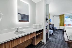 A modern hotel room with a large bathroom vanity, a bed, a desk, and a view of the city.