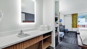 A modern hotel room with a white vanity, a large mirror, a wooden cabinet, a bed, a desk, and a view of the city through the window.