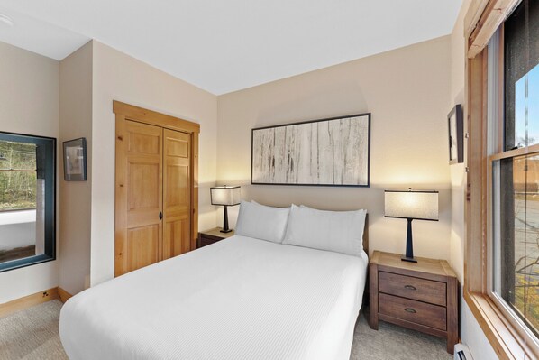 A bedroom with a large bed, two bedside tables with lamps, a wooden wardrobe, a framed artwork on the wall, and a window with a view. - Charming Retreat in Keystone w/ Private Patio & Fireplace, Close to River Run (Keystone)