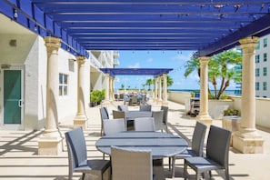 An outdoor patio with round tables and chairs, covered by a blue canopy, and surrounded by columns.