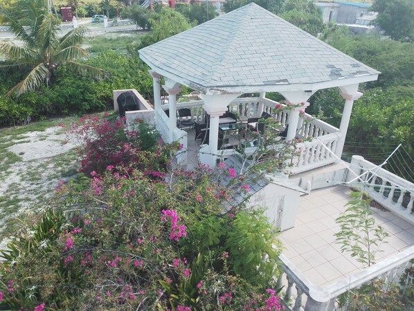 Ein Gartenpavillon mit einem Ziegeldach, umgeben von üppigem Grün und rosa Blüten.
