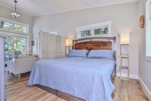 Un dormitorio con una cama grande, cabecera de madera, dos mesitas de noche con lámparas, una ventana con vistas a árboles y un balcón con mobiliario exterior.