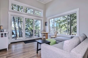 A spacious living room with large windows, a grey sofa, a small table, and a chair.