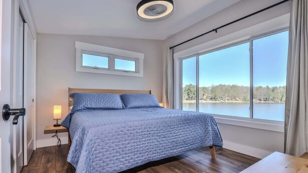 Una camera da letto con una grande finestra, un letto con biancheria azzurra, una lampada da comodino e un pavimento in legno.