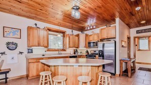 A modern kitchen with wooden cabinets, a central island with stools, stainless steel appliances, and a large window. - Eikedahls' Shoreview Lake House (Minocqua)