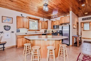 A modern kitchen with wooden cabinets, a central island with stools, stainless steel appliances, and a large window. - Eikedahls' Shoreview Lake House (Minocqua)