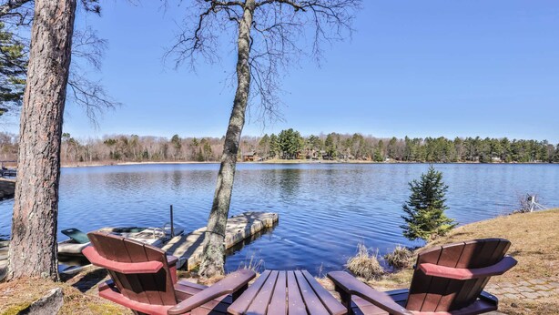 A lakeside picnic area with two Adirondack chairs and a wooden table, surrounded by trees and a clear view of the lake. - Eikedahls' Shoreview Lake House (Minocqua)