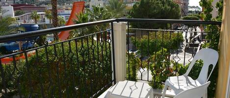 Un balcon avec des chaises et une table en plastique blanc, d’où l’on peut admirer un paysage urbain composé d’immeubles et d’espaces verts.