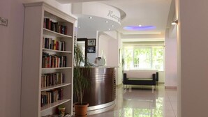 A reception area with a curved reception desk, a bookshelf filled with books, a potted plant, and a bench. - Kleopatra Hotel (Alanya)