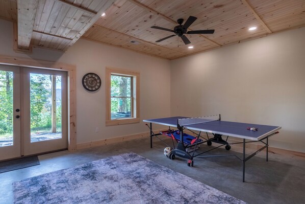 A room with a ping pong table, a ceiling fan, and a large window with a view of trees. - Summit View Lodge - Hot Tub - WIFI (Whittier)