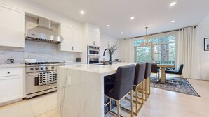 Een moderne keuken met witte kasten, een centraal eiland en een eetgedeelte met uitzicht op bomen.