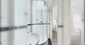 A modern bathroom with a glass-enclosed shower, a white sink, and a bed visible through an open door.