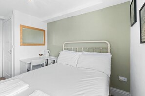 A bedroom with a bed, a bedside table, a chair, a desk, a mirror, and a wall socket. - Charming Seaside Room | Steps from Brighton Pier (Brighton and Hove)