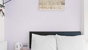 Una cama con almohadas blancas, una mesita de noche de madera con una tetera y botellas de agua, y un cuadro colgado en la pared con botellas.
