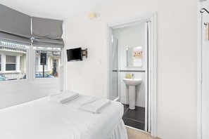 A hotel room with a bed, a TV mounted on the wall, a bathroom with a sink and mirror, and a view of the outside through a window with blinds.