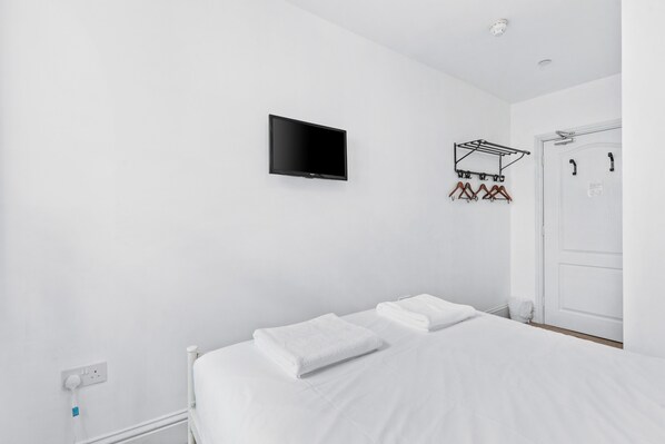 A minimalist hotel room with a bed, a flat-screen TV mounted on the wall, a coat rack with hangers, and a door with a handle.