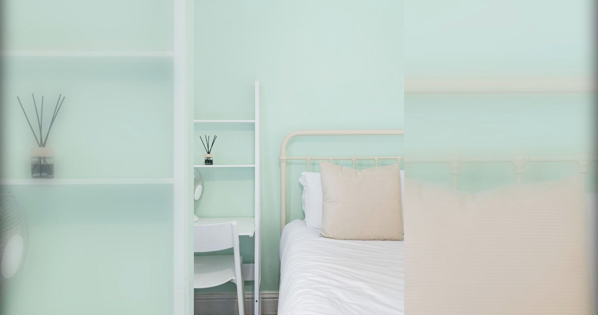 Une chambre à coucher avec un lit, une table de chevet blanche et une étagère sur laquelle se trouvent un diffuseur et un petit miroir.