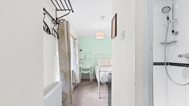 Un couloir étroit mène à une salle de bain équipée d’une douche, d’un radiateur et d’un lit avec une table de chevet.
