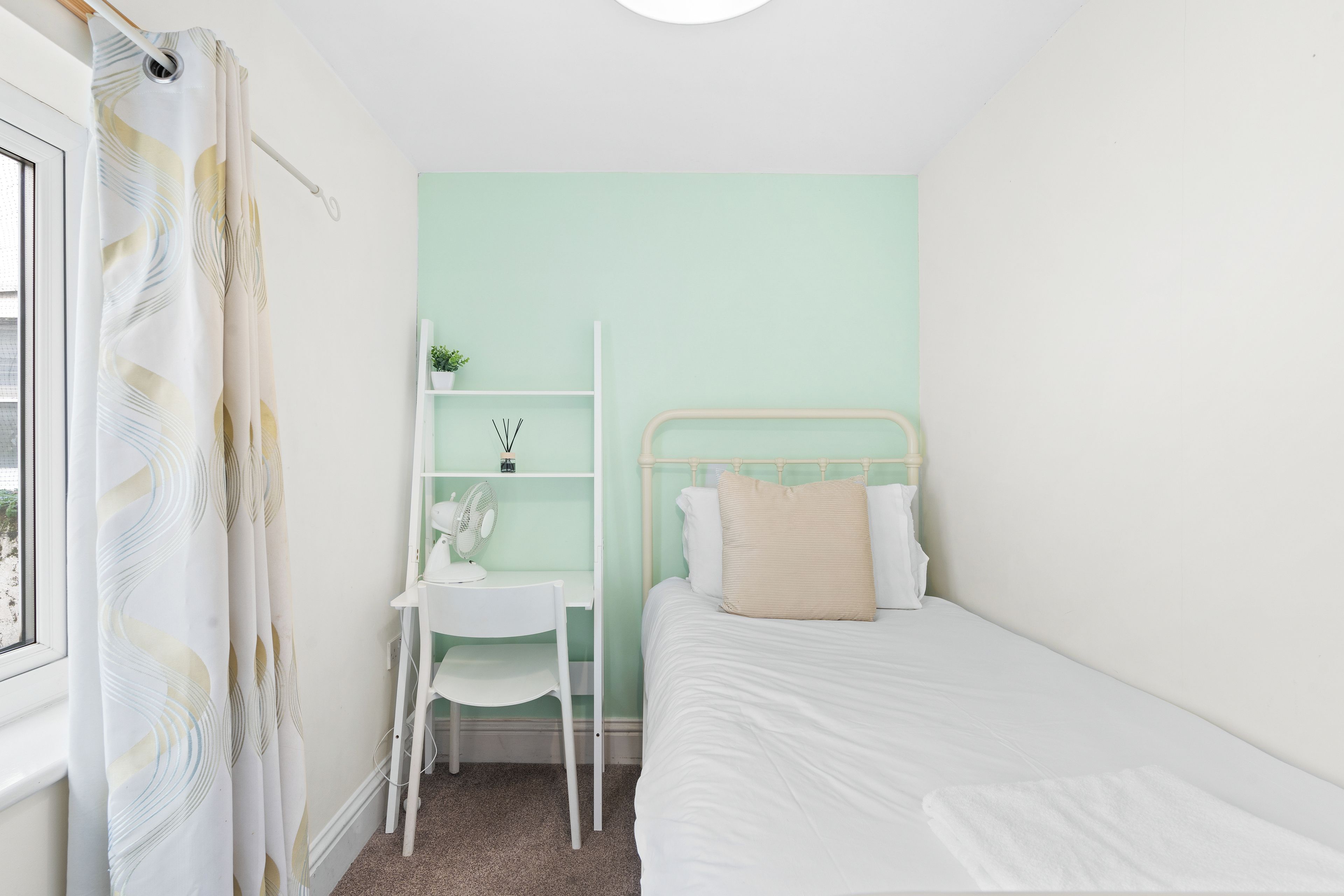 A bedroom with a bed, a desk with a chair, a shelf with a plant, and a window with curtains.