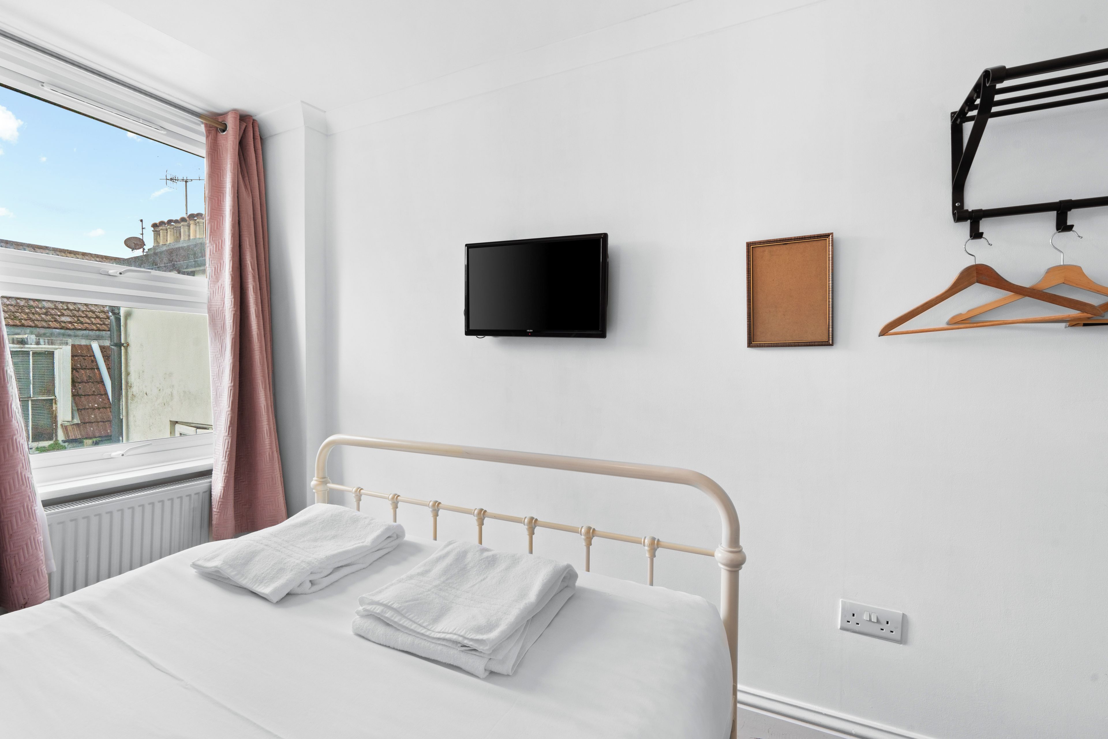 Un dormitorio con una cama, un televisor de pantalla plana, un perchero con ganchos y una ventana con cortinas.