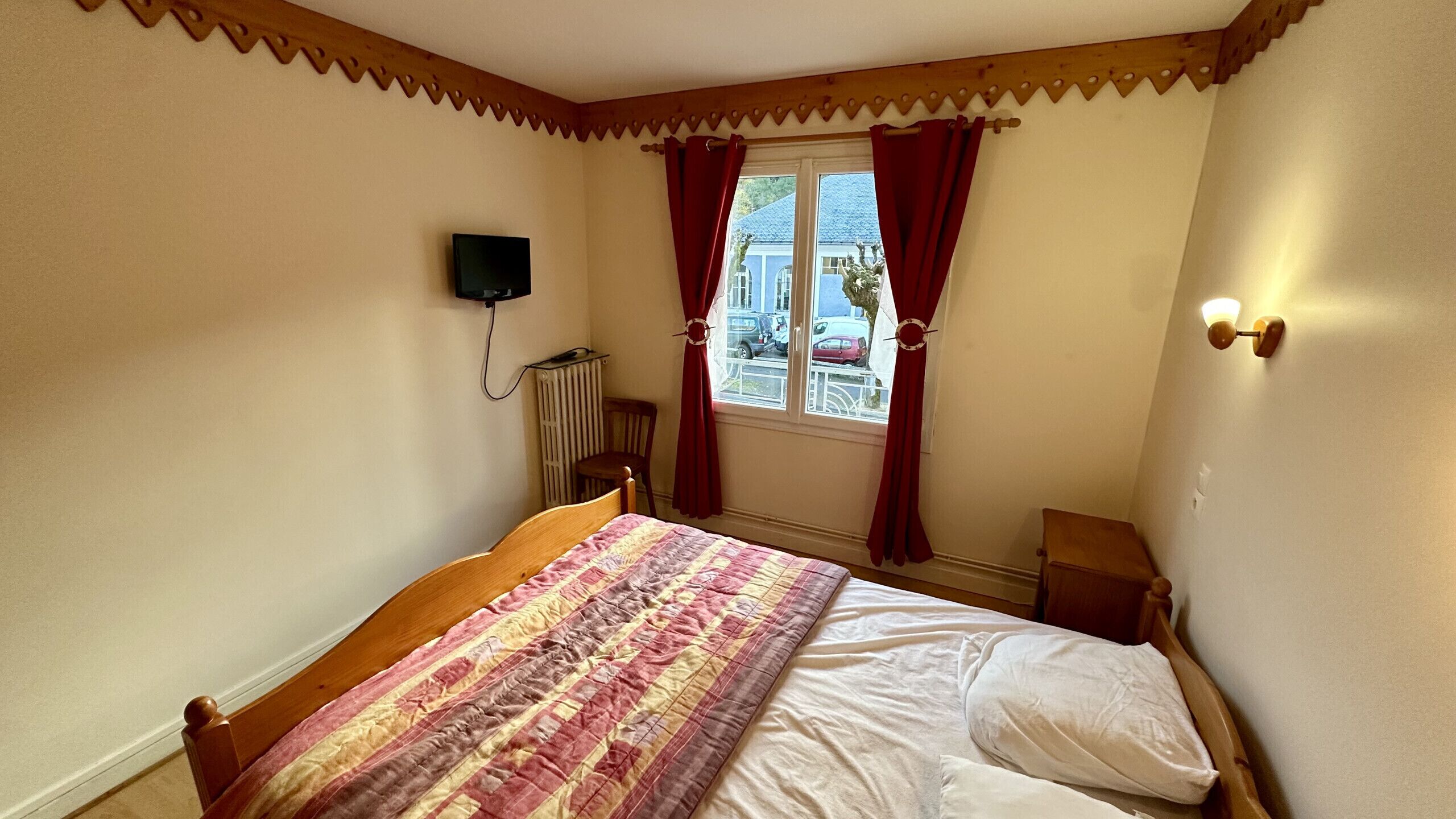 Ein Schlafzimmer mit einem hölzernen Bett, einem Heizkörper, einem Fenster mit roten Vorhängen und einer Wandleuchte.