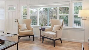 Een kamer met twee beige fauteuils, een tafeltje en een lamp. Er zijn grote ramen met zonwering, een deur en een houten vloer.